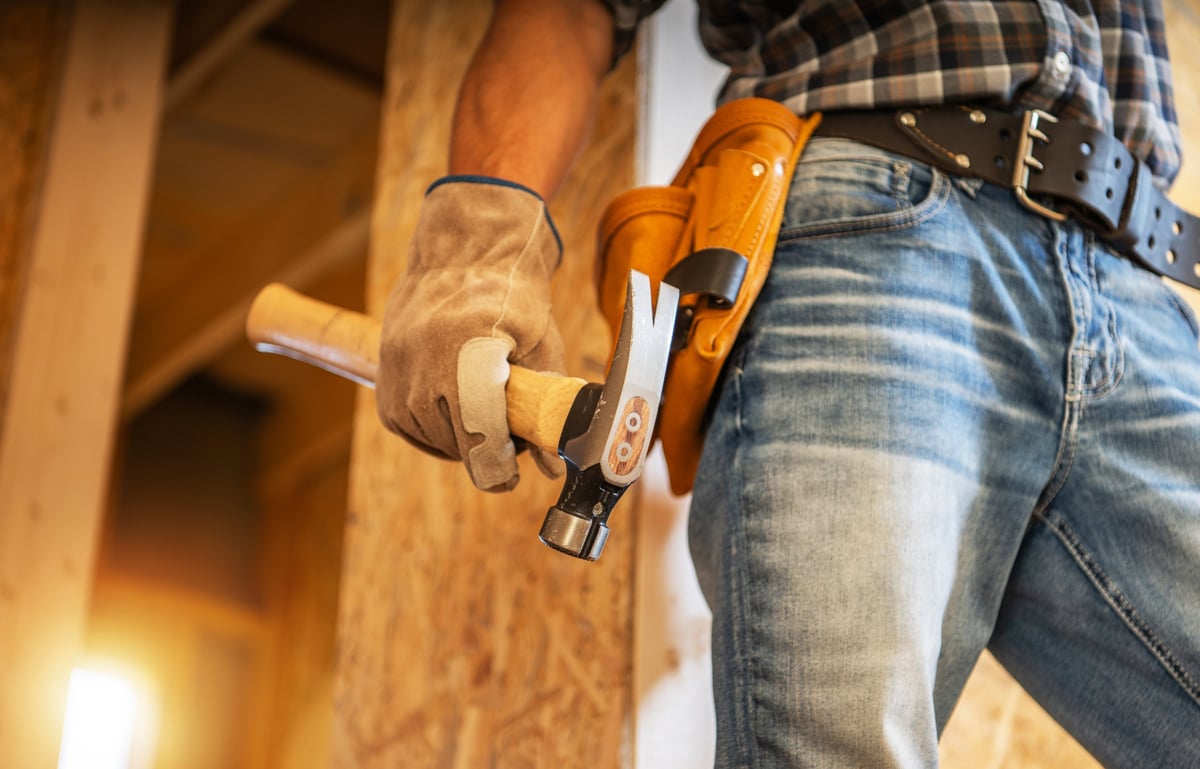 A skilled carpenter holds a hammer while wearing gloves and a tool belt, standing in a construction area illuminated by warm evening light.