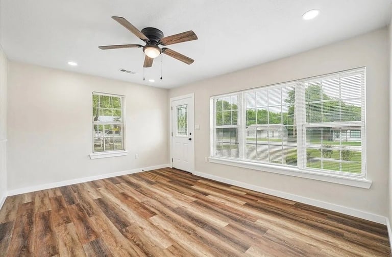 Empty room with wood flooring, ceiling fan, and large windows overlooking green landscape