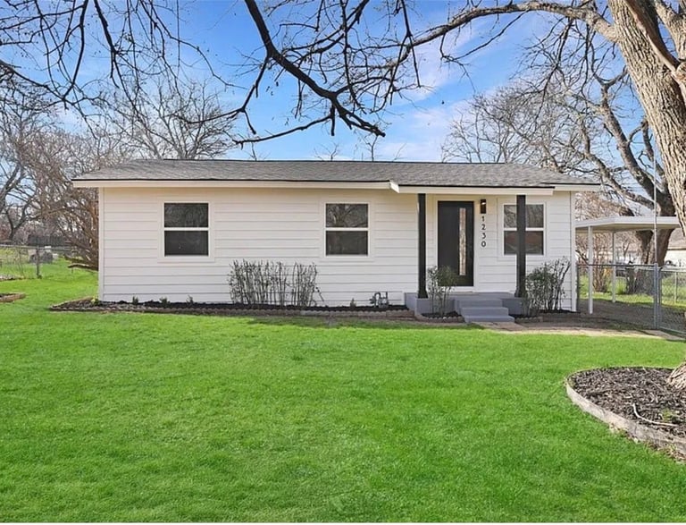 Single-story white house with dark roof on green lawn, bare trees in background, black door and shutters, landscaping bed visible