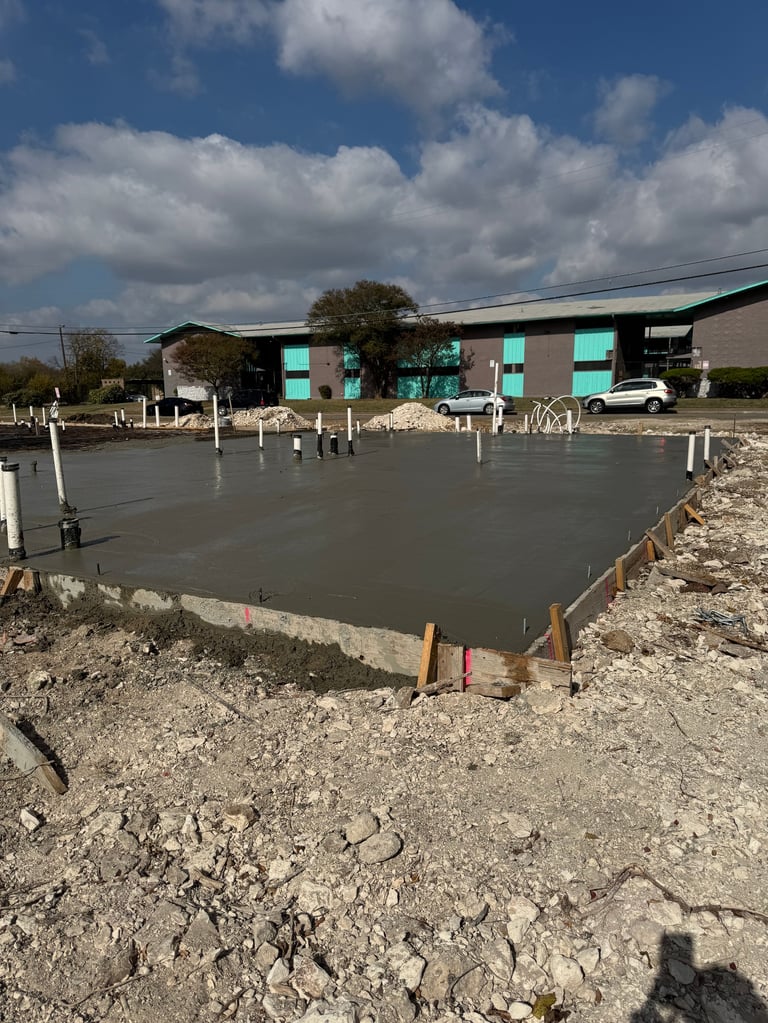 Construction site with freshly poured concrete foundation and turquoise building structure in background under blue sky