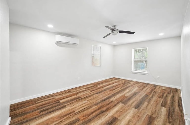 Empty room with wood-grain flooring, white walls, ceiling fan, air conditioning unit, and two windows with natural light