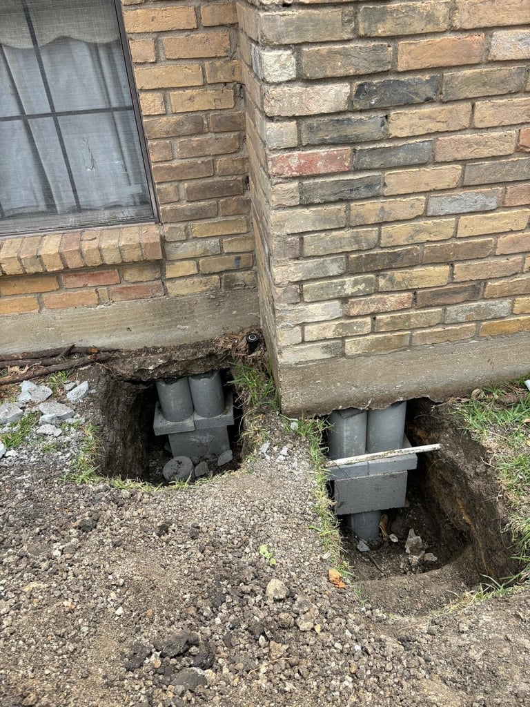 Two black metal drainage pipes or downspouts installed at the base of a brick building foundation against soil and grass