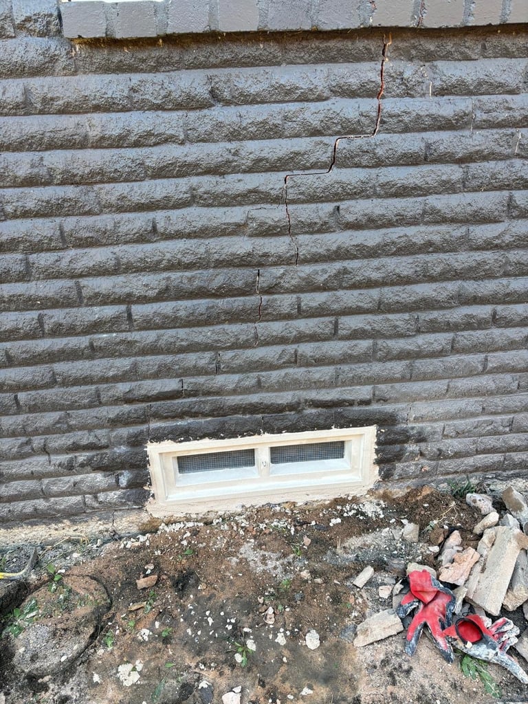 White basement window set in a weathered concrete brick wall with soil and debris below