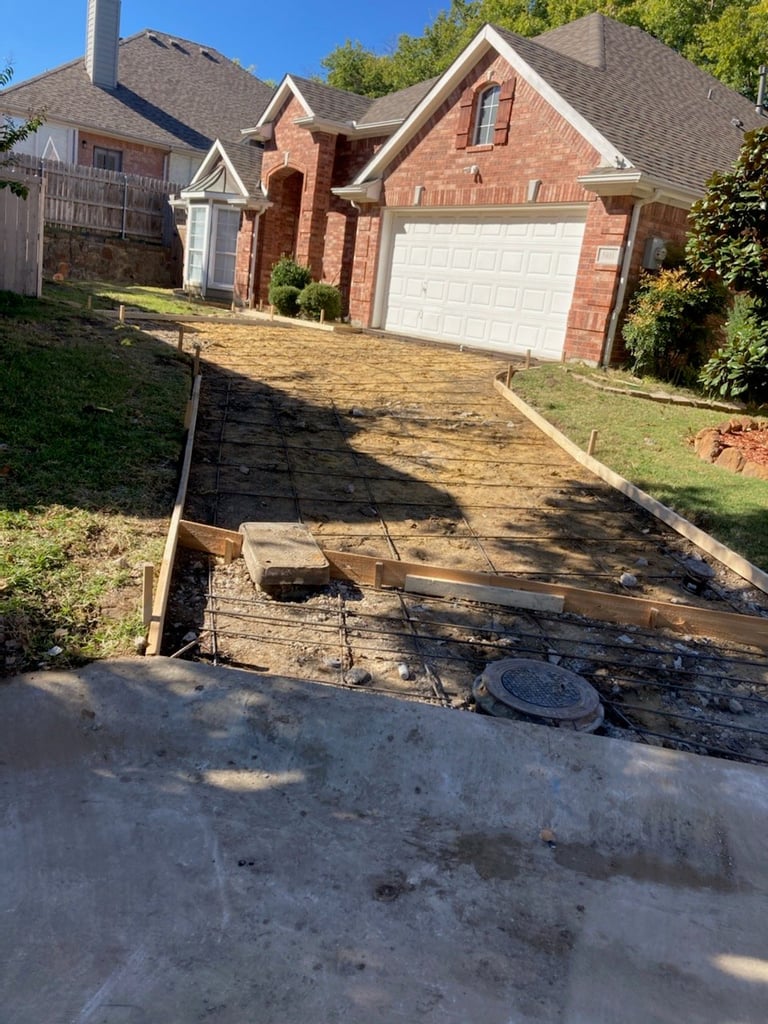 Residential driveway under construction with wooden forms and metal drainage pipe in front of a brick two-story house