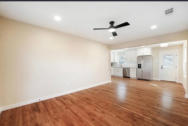 Empty living room with wood flooring, beige walls, ceiling fan, recessed lighting, and kitchen visible through doorway