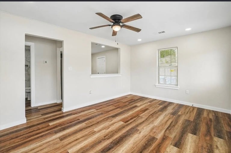Empty bedroom with wood flooring, ceiling fan, built-in shelving, doorway, and windows providing natural light